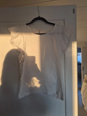 Vit topp med spetsdetaljer - Söt vit topp med korta ärmar och spetsdetaljer längs axlarna. Rund halsringning och rak passform. Perfekt att matcha med jeans eller kjol för en fräsch look.