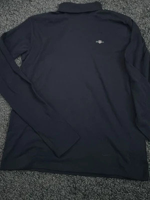 Mörkblå långärmad skjorta från Gant - Klassisk mörkblå långärmad skjorta från Gant med broderad logga på bröstet. Skjortan har en enkel och stilren design med krage och är tillverkad i mjuk bomull som känns skön mot huden. Perfekt för en clean och avslappnad look.