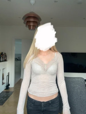 Beige långärmad intimissimi med spets - Snygg beige långärmad topp med v-ringning och spetsdetaljer vid halsen. Modellen är croppad och har en feminin passform som framhäver figuren. Perfekt att styla med jeans eller kjol för en trendig look.