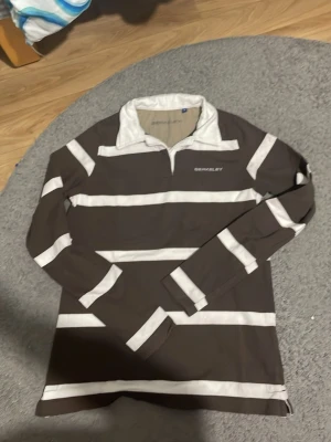 Randig långärmad skjorta från Berkeley - Säljer en brun och vit randig långärmad skjorta från Berkeley. Skjortan har en klassisk krage, breda horisontella ränder och en broderad logga på bröstet. Tillverkad i mjuk bomull och perfekt för en avslappnad stil.