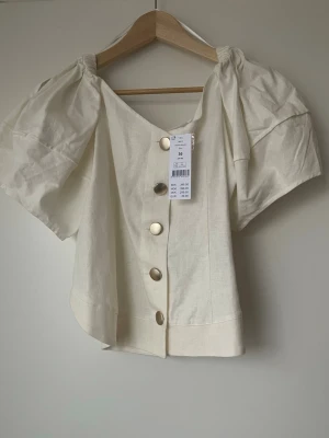 Krämvit blus med puffärm och knappar - Säljer en krämvit blus från H&M med puffiga korta ärmar och stora guldfärgade knappar framtill. Toppen har en v-ringad hals och är tillverkad i ett linneliknande material som känns luftigt och somrigt. Perfekt för dig som gillar trendiga detaljer och en avslappnad look.