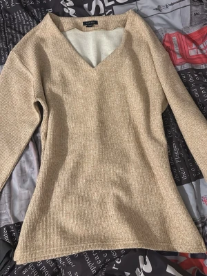 Beige stickad tröja med v-ringning - Beige stickad tröja från AMISU med v-ringad hals och långa ärmar. Tröjan har en mjuk, ribbad struktur och är lätt figursydd för en snygg passform. Perfekt för lager på lager och enkel att matcha med olika stilar.