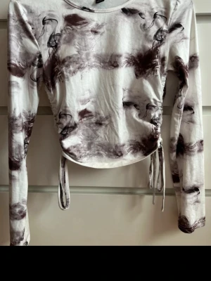 Vit och brun långärmad topp med snörning - Trendig långärmad topp i vitt med abstrakt brunt mönster. Toppen har snörning i sidorna för justerbar passform och är croppad. Tillverkad i mjuk bomullsmix, perfekt för en cool och avslappnad stil.