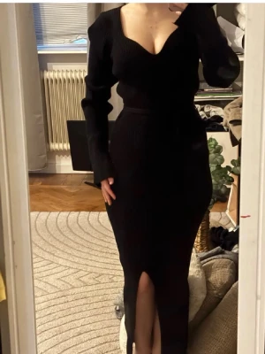 Svart ribbad långklänning med slits - Snygg svart långklänning i ribbat material med djup urringning och långa ärmar. Klänningen har en figurnära passform och en hög slits framtill och knyt på midjan som ger en trendig look. Perfekt för dig som gillar stilrena och moderna plagg.