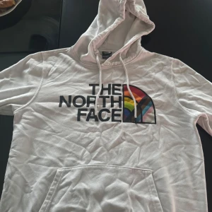 Vit hoodie från The North Face - Säljer en vit hoodie från The North Face med regnbågsfärgad logga på bröstet. Den har en klassisk känguruficka och justerbar huva med dragsko. Perfekt för en avslappnad stil.