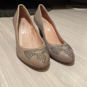 Beige klackskor från MJUS - Snygga beige ballerinaskor från Mjus med dekorativa nitar i form av en rosett på tån. Skorna har en låg klack och är perfekta för en stilren look. De är i storlek 37 och har en elegant design som passar till många tillfällen.