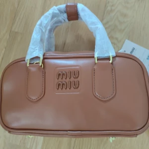Brun handväska från Miu Miu - Snygg brun handväska från Miu Miu i skinn med guldfärgade detaljer. Väskan har en praktisk dragkedja upptill och ett elegant Miu Miu-märke på framsidan. Perfekt för att ge din outfit en lyxig touch.
