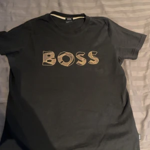 Svart t-shirt från Boss - Snygg svart t-shirt från Boss med ett stort, stilrent tryck på framsidan. Tillverkad i mjuk bomull för en bekväm passform. Perfekt för en casual look.