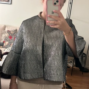 Silverfärgad jacka från Kappahl - Säljer en cool silverfärgad jacka från Kappahl med en unik textur. Jackan har trekvartsärmar och en öppen design med små knäppen.