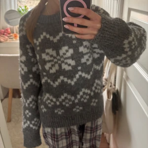 Grå stickad tröja med snöflingemönster - Mysig grå stickad tröja med vitt snöflingemönster. Perfekt för kyliga dagar med långa ärmar och ribbstickade kanter. En klassisk och stilren design som passar till mycket.