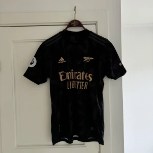 Arsenal matchtröja - Snygg svart fotbollströja från Adidas med guldiga detaljer och korta ärmar. Tröjan har en diskret mönstring och Arsenal loggan på bröstet.