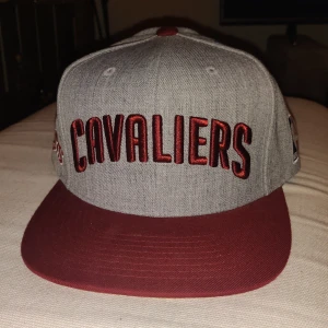 Grå och vinröd Cavaliers keps (snapback)från Mitchell & Ness - Snygg keps i grått med vinröd skärm och broderad Cavaliers-logga. Kepsen har flera broderade detaljer och justerbar passform. Perfekt för basketfans! (Kepsen är aldrig använd bara legat i ett skåp dock är klisterlapparna på skärmen borttagna)
