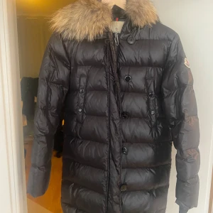 Svart dunjacka från Moncler - Säljer en svart dunjacka från Moncler med en lyxig pälsdetalj på huvan. Jackan har både dragkedja och knappar framtill samt Monclers logga på ärmen. Perfekt för kalla vinterdagar. Jackan är äkta, jag jobbar som flytt och fick jackan av en jätterik kund som tänkte slänga den. Pris kan möjligtvis diskuteras lite, i så fall vid snabb affär eller liknande.