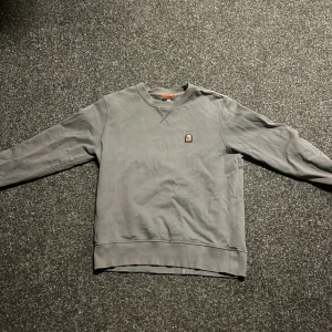 Grå sweatshirt från Parajumpers - Säljer en stilren grå sweatshirt från Parajumpers i fleece. Tröjan har en liten broderad logga på bröstet och ribbade muddar vid ärmslut och nederkant. Perfekt för en avslappnad look.