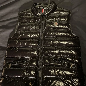 Svart glansig dunväst från Moncler - Snygg svart glansig dunväst från Moncler med dragkedja som är lite trasig i botten och två fickor framtill. Perfekt för att hålla värmen med stil. Den har en klassisk Moncler-logga på bröstet och en unik serietidningsetikett på insidan som sitter löst. Inte 100 på om den är äkta där av priset