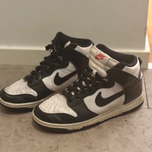 Svartvita Nike Dunk sneakers - Snygga svartvita Nike Dunk sneakers med klassisk design. Skorna har en hög modell med snörning och den ikoniska Nike-loggan på sidan. Perfekta för en stilren look.