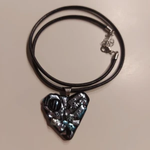 Blått hjärtformat halsband i glas - Unikt blått halsband med ett hjärtformat hänge som är gjort av glas. Hänget är fäst på en svart rem med justerbar kedja. Perfekt för att ge en edgy touch till din stil. Skriv om ni har någon fundering!✨️