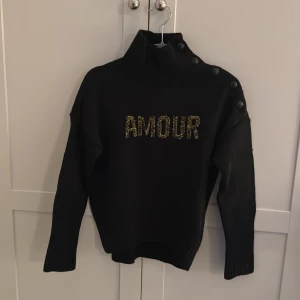 Svart tröja med text från Zadig & Voltaire - Snygg svart tröja från Zadig & Voltaire med texten 'AMOUR' i glittriga bokstäver på framsidan. Storleken på tröjan är 10 år 138. Tröjan har en hög krage och knappar längs axeln. Den här tröjan är för liten på mig och har inte används så mycket tröjan kommer även med en extra knapp på lappen inuti tröjan