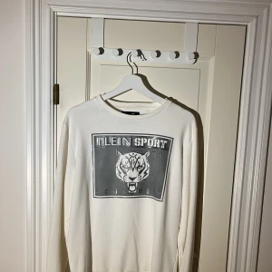 Vit långärmad tröja från Plein Sport - Säljer en stilren vit långärmad tröja från Plein Sport med ett stort tryck av en tiger och text på framsidan. Perfekt för en sportig look. Tröjan har ribbade muddar vid ärmslut och nederkant. Tröjan är köpt från johnells.se nypris (3890kr). Har du minsta lilla fundering så hör av dig! Tröjan är som ny!