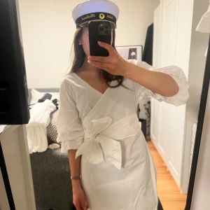 Vit omlottklänning med knyt - Säljer en stilren vit omlottklänning med knyt i midjan. Klänningen har en v-ringad design och trekvartsärmar, perfekt för sommarens festligheter. Dem funkar även som en oneshoulder klänning med. Perfekt studentklänning!! Finns ett litet inhopp sytt hål i armhålan som knappt syns annars i mycket bra skick.❤️