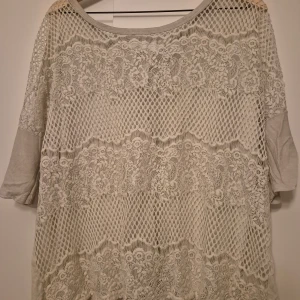 Grå topp med spetsdetaljer - Säljer en unik grå topp med spetsdetaljer och nätmönster. Toppen har trekvartsärmar och en lös passform, perfekt för en stilren look. Den är tillverkad i ett mjukt material som ger en bekväm känsla. Passar bra till både jeans och kjol.
