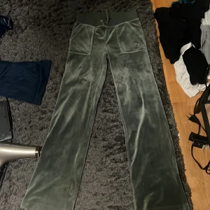 Gröna velourbyxor från Juicy Couture, använda fåtal gånger och prisvä kan diskuteras - Snygga gröna velourbyxor från Juicy Couture med bred resår i midjan och snörning. Byxorna har fickor med knappar både fram och bak, vilket ger en cool look. Perfekta för en avslappnad stil.