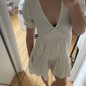 Vit omlottklänning - Säljer en söt vit omlottklänning med korta puffärmar och knytband i midjan. Klänningen har en volangdetalj vid fållen och en v-ringad hals. Perfekt för sommarens varma dagar! Student 