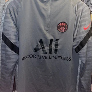 Grå träningströja - Snygg grå träningsjacka från Nike med PSG-logga på bröstet och röd Nike-swoosh på ärmen. Tröjan har en halv dragkedja och svarta detaljer på ärmarna. Perfekt för fotbollsträning eller vardagsbruk.