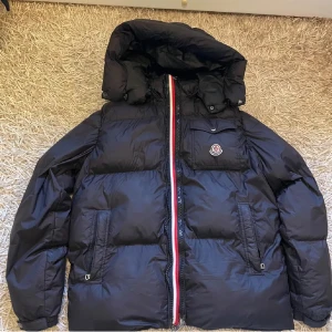 Svart dunjacka från Moncler - Snygg svart dunjacka från Moncler med huva och dragkedja i rött, vitt och blått. Jackan har en klassisk pufferdesign och en praktisk bröstficka med Moncler-logga. Perfekt för kyliga dagar. Köpte för 20tusen.