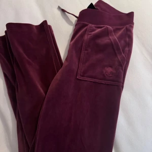Vinröda mjukisbyxor från Juicy Couture - Snygga vinröda mjukisbyxor från Juicy Couture i mjukt sammetstyg. Byxorna har en praktisk ficka med knappar och en broderad logga. Perfekta för en avslappnad stil.