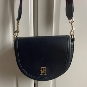 Mörkblå axelväska från Tommy Hilfiger  - Snygg svart axelväska i skinn med en elegant logotypdetalj i guld på framsidan. Väskan har en justerbar axelrem med guldfärgade spännen. Aldrig använd 