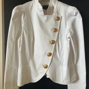 Vit kavaj från Gare De Robe - Snygg vit kavaj från Gare De Robe med unika guldknappar i en asymmetrisk design. Kavajen har långa ärmar och en stilren look som passar perfekt för en elegant touch. Perfekt för dig som vill sticka ut med stil.