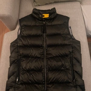 Mörk grön  dunväst från Parajumpers - Säljer en snygg mörk grön  dunväst från Parajumpers med dragkedja och hög krage. Perfekt för kyliga dagar när du vill hålla värmen utan att kompromissa med stilen. Västen har en cool detalj med en gul knapp vid halsen.