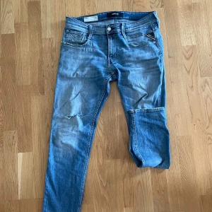 Blå jeans från Replay - Snygga blå jeans från Replay med klassisk femficksdesign och knappgylf. De har en lätt slitning på framsidan och en läderdetalj vid midjan. Perfekta för en avslappnad stil.
