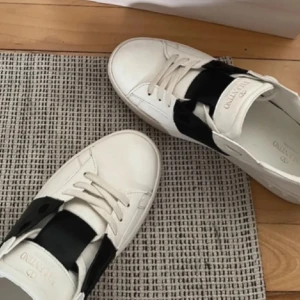 Valentino Garavani sneakers - Snygga Valentino Garavani sneakers i vitt med en bred svart rem över snörningen. Skorna har en stilren design med Valention-logga på plösen. Perfekta för en trendig look. 