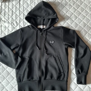 Svart hoodie från Comme des Garçons, liten i storleken. - Säljer en stilren svart hoodie från Comme des Garçons med dragkedja och huva. Storlek S men liten i storleken. Sparsamt använd i mycket bra skick. Nypris 2700kr