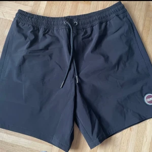 Svarta badshorts från Colmar - Snygga svarta badshorts från italienska Colmar med elastisk midja och snörning för perfekt passform. Shortsen har en bakficka med knapp och Colmar-logga på framsidan. Perfekta för sommarens alla äventyr.