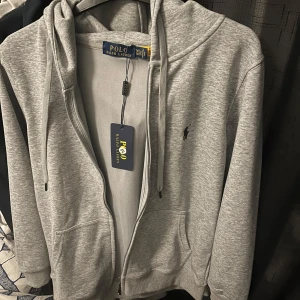 Grå hoodie från Ralph Lauren - Säljer en stilren grå hoodie från Ralph Lauren med dragkedja och justerbar huva. Tröjan har en liten broderad logga på bröstet och är tillverkad i mjukt material för extra komfort. Perfekt för en avslappnad look.