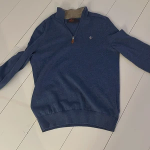 Blå tröja från Morris - Säljer en stilren blå tröja från Morris i 100% merinoull. Tröjan har en dragkedja vid halsen och ett litet broderat emblem på bröstet. Perfekt för kyligare dagar. Bra skick och storlek s vuxen