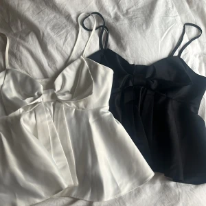 Svart och vit topp med rosett - Två eleganta toppar i svart och vit färg med tunna axelband och stor rosett framtill. Topparna har en lätt utsvängd passform och är tillverkade i ett glansigt material som ger en lyxig känsla.