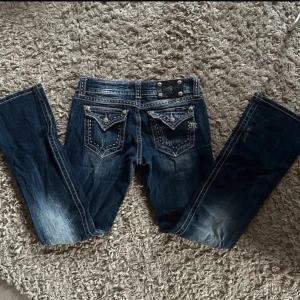 Blå jeans från Miss Me - Snygga blå jeans från Miss Me med dekorativa bakfickor och slitna detaljer. Jeansen har en låg midja och en bootcut-stil. De är prydda med knappar och broderade detaljer som ger en unik touch. Bootcut varianten är väldigt svårt att få tag i😇💘Sitter som en s och lite mot xs❤️