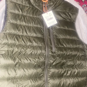 Olivgrön dunväst från Swix - Säljer en olivgrön dunväst från Swix i storlek XL. Västen har en dragkedja framtill och är perfekt för kyligare dagar. Den är lätt och smidig att bära, vilket gör den till ett bra val för utomhusaktiviteter.