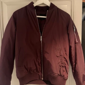Vinröd bomberjacka  - Säljer en snygg vinröd bomberjacka från New Look. Jackan har en dragkedja framtill och en ficka med dragkedja på ärmen. Den är tillverkad i ett glansigt material och har ribbade muddar vid ärmslut och nederkant. Perfekt nu till våren! Den är i storlek 6 vilket jag tror är samma som S i EU storlek, passar mig som vanligtvis bär S!