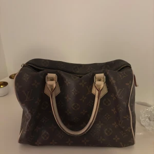 Brun handväska från Louis Vuitton - Säljer en elegant brun handväska från Louis Vuitton med det klassiska monogrammönstret. Väskan har två handtag och en dragkedja upptill som dessvärre är trasig (se sista bilden) därav priset, väskan går fortfarande att stänga. Perfekt för att ge en lyxig touch till din outfit.