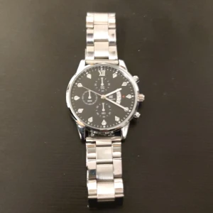 Silverfärgad klocka med svart urtavla - Stilren klocka med silverfärgad metallarmband och svart urtavla. Klockan har romerska siffror och flera små urtavlor för extra funktioner. Perfekt för den som gillar en klassisk och elegant stil.