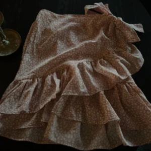 Rosa volangkjol med knytband - Söt rosa kjol med volanger och knytband i midjan. Kjolen har ett diskret mönster och är perfekt för en somrig look. Den är kort och har en luftig passform.