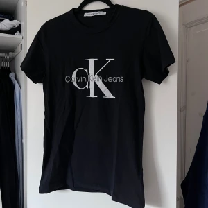 Svart t-shirt från Calvin Klein Jeans - Snygg svart t-shirt från Calvin Klein Jeans med det ikoniska CK-logotypen tryckt i vitt på framsidan. Perfekt för en stilren och avslappnad look. T-shirten har en klassisk passform och korta ärmar. Använd max 3 gånger. Önskar du veta exakta mått? Tveka inte att höra av dig!