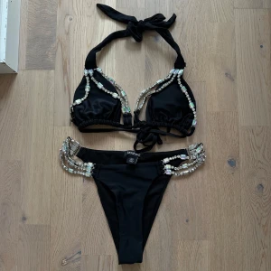 Svart bikini med strassdetaljer från Boohoo - Svart bikini från Boohoo med glittrande strassdetaljer längs kanterna. Toppen har knytband i nacken och ryggen för justerbar passform. Bikinitrosorna har dekorativa paljetter på sidorna som ger en extra touch av glamour.