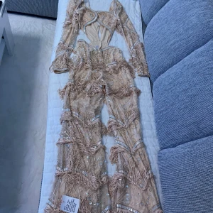 ASOS LUXE - ljusrosa jumpsuite - Säljer en unik beige oneshoulder klänning med fransar och paljettdetaljer. Klänningen har en öppen rygg och är dekorerad med glittriga element som ger en festlig känsla. Perfekt för en speciell kväll!