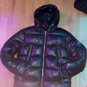 Svart pufferjacka från Supply & Demand - Säljer en svart pufferjacka från Supply & Demand med en glansig finish. Jackan har en dragkedja framtill och en huva för extra skydd. Perfekt för kyliga dagar med sin vadderade design. Märkeslogga på ärmen ger en cool touch. Priset är inte hugget i sten